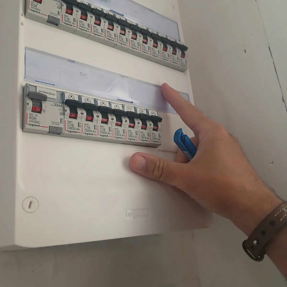 Main ouvrant le capot d’un tableau électrique, rangées de disjoncteurs