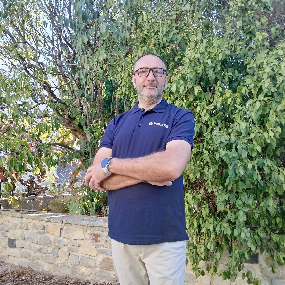 Guillaume Pons, bras croisés, devant un muret de pierres et une haie